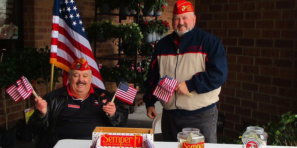 Two members in dress blues beside the Marine Corps flag and the American flag